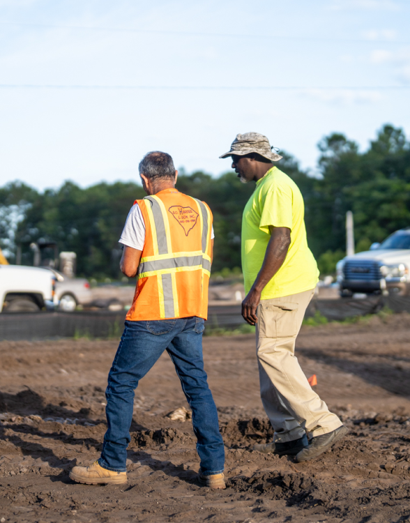 Two workers talk on a dirt site with vehicles behind.
