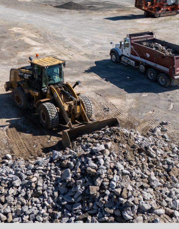 Front loader moves rubble; dump truck waits nearby onsite.
