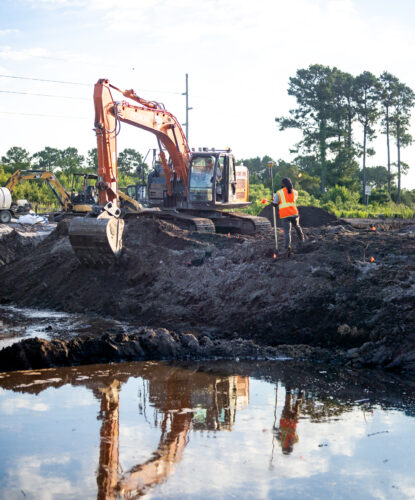 Excavator and worker in vest on muddy site near trees.
