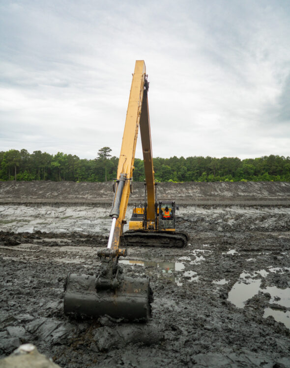 Long-arm excavator digging mud at site, trees behind.