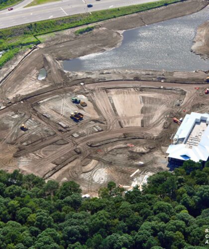Aerial view: construction site, vehicles, equipment, road, pond, trees.