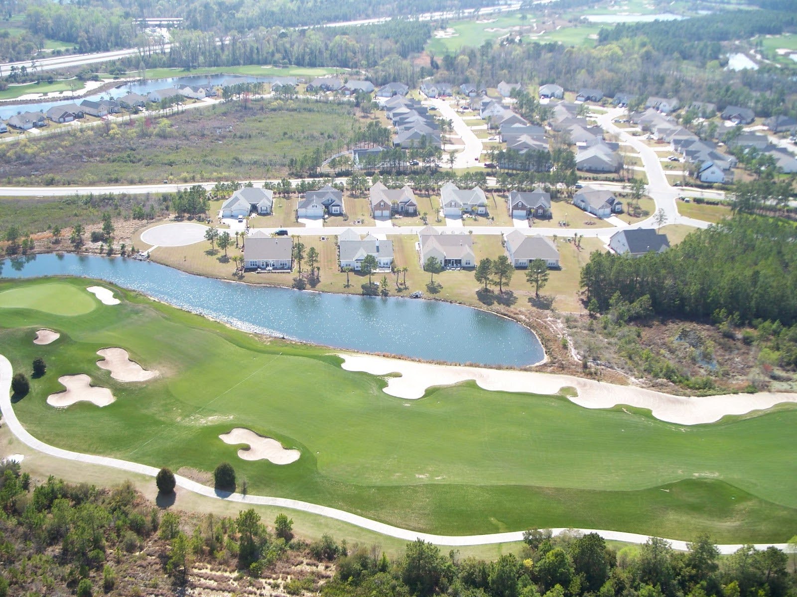 Aerial view: golf course, sand traps, pond, houses, tree-lined streets.