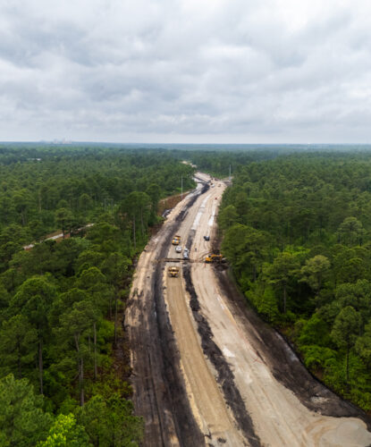 Highway construction cuts through dense forest, vehicles visible below.