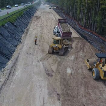 Workers, dump truck, and steamroller at construction site by highway.