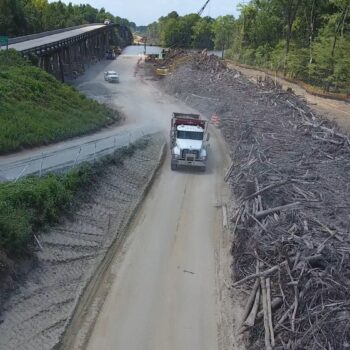 White dump truck drives by logs, debris, trees, overpass.