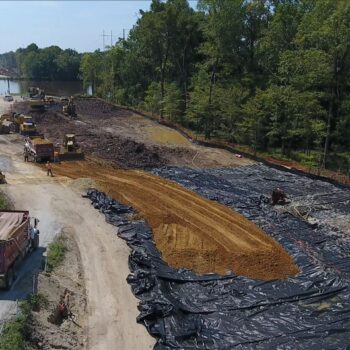 Aerial view: road construction site with machinery, plastic sheeting, trees.