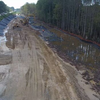 Construction site by highway: machinery, dirt, water, trees, barriers.