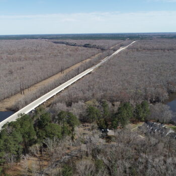 A straight bridge crosses river and leafless forest under clear sky.
