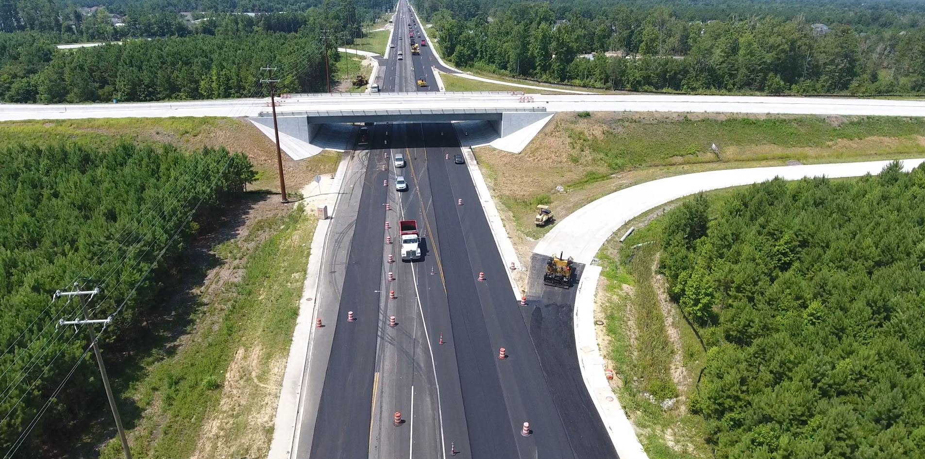 Highway roadwork with cones, workers, cars near overpass, greenery.