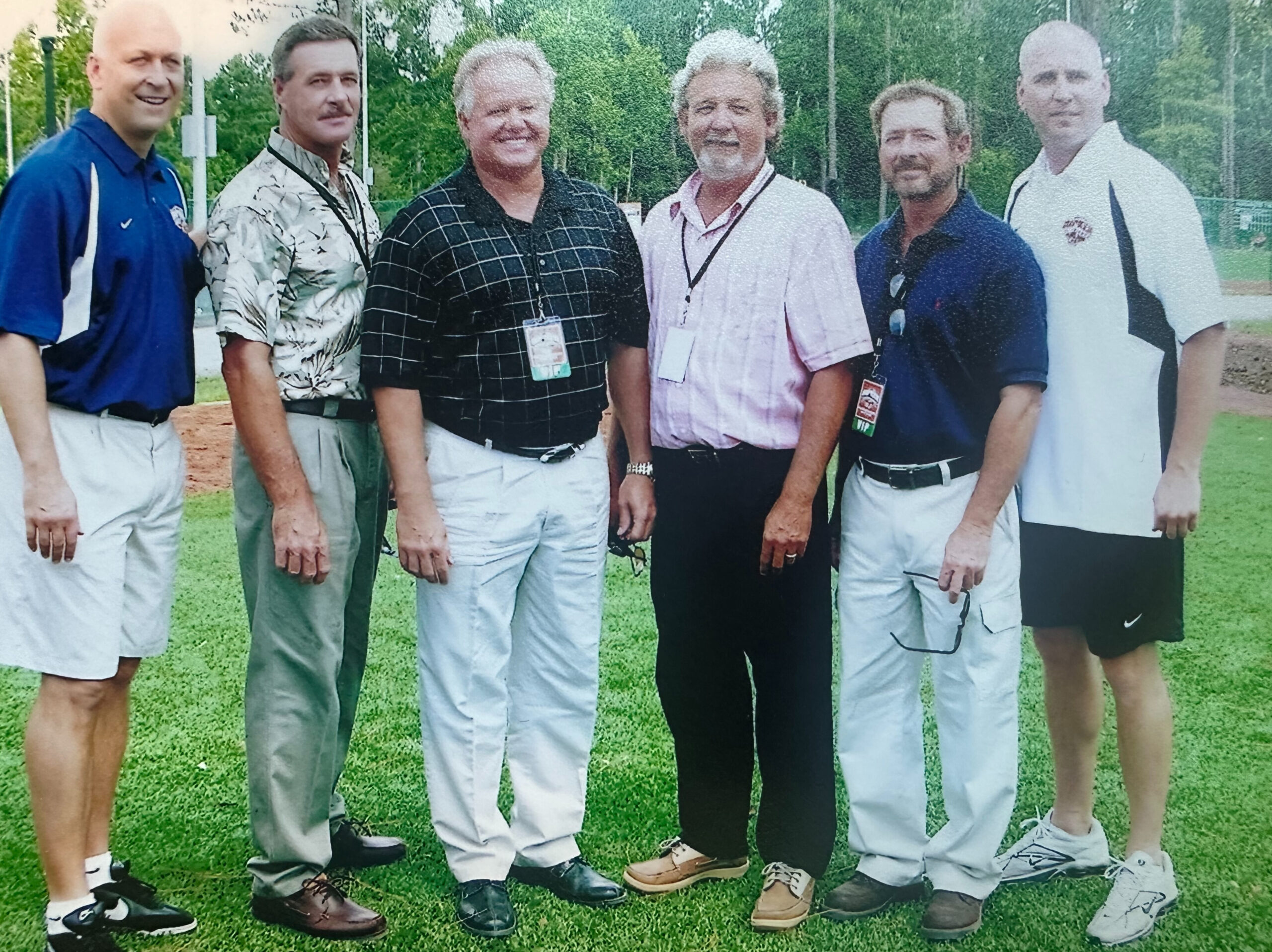 Six men pose on grass with trees in the background.