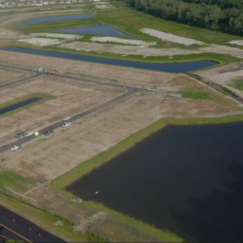 Aerial view: construction site, roads, ponds, vehicles, trees, homes.