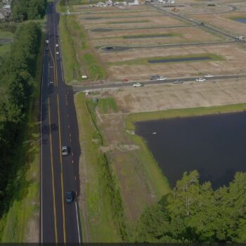 Aerial view: houses, main road, vacant lots, pond in daylight.