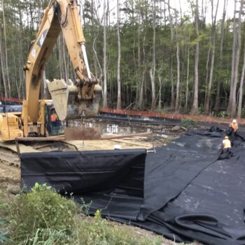 Excavator and workers install black sheets by wooded site.