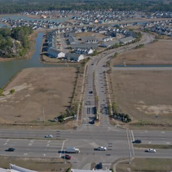 Suburban aerial: main road, houses, pond left, cars visible.