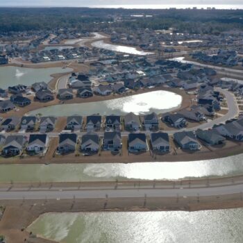 Aerial view: suburban homes, winding roads, lakes, clear skies.