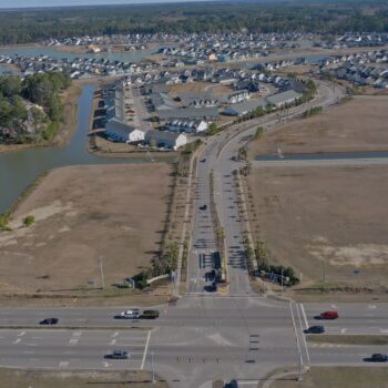 Aerial view: gated community entrance, roads, houses, pond, cars.