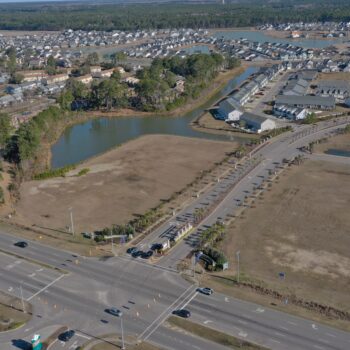 Aerial view: suburb, pond, main road, roundabout, trees beyond.