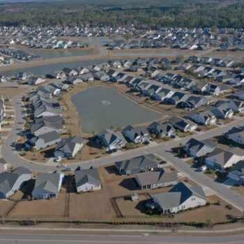 Aerial view: suburban homes, curved roads around pond with fountain.