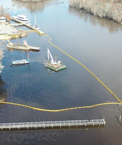 Aerial view: river, barges, yellow barriers, dock, wooded shore.