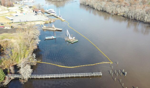 Aerial view: river, barges, yellow barriers, dock, wooded shore.
