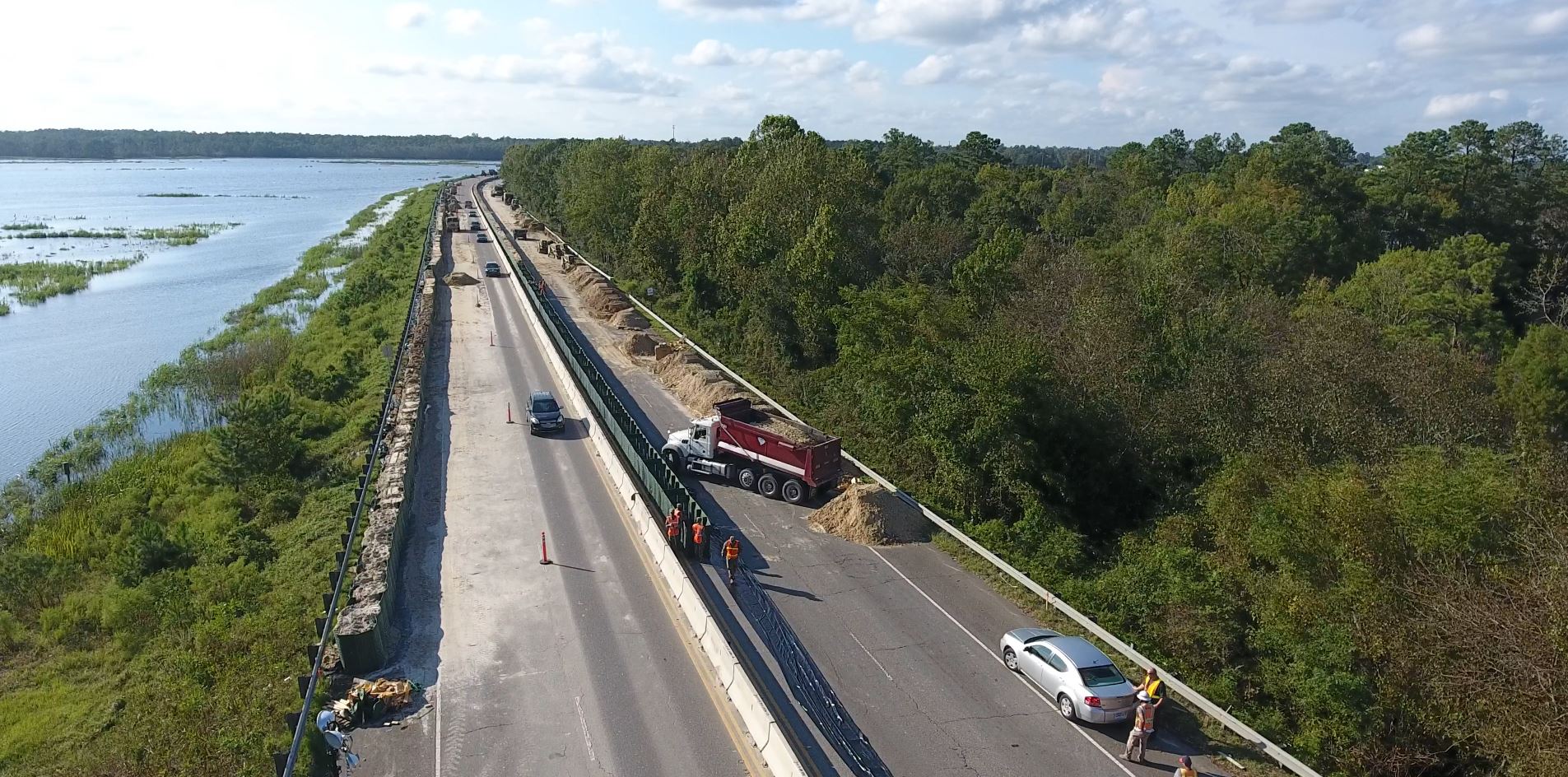 Aerial view: road construction, vehicles, workers by water and woods.