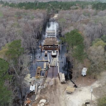 Aerial view: bridge construction in swampy woods, house nearby.
