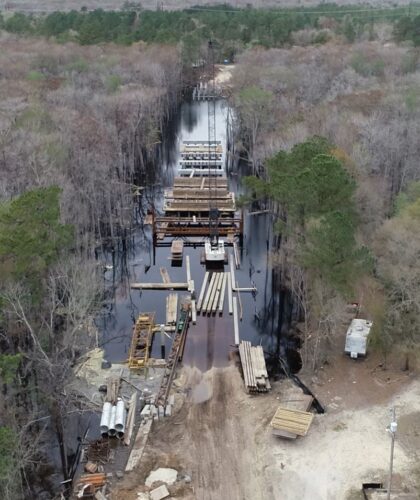 Aerial view: bridge construction in swampy woods, house nearby.
