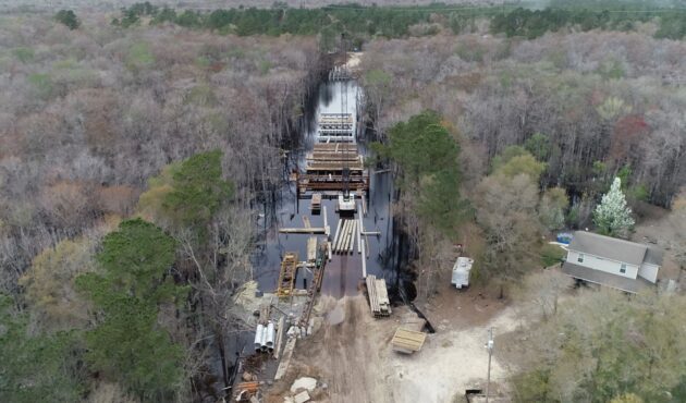 Aerial view: bridge construction in swampy woods, house nearby.
