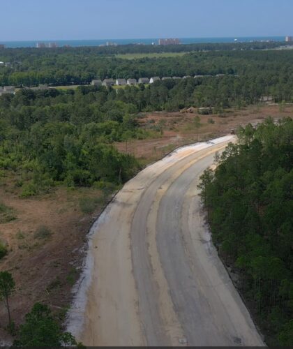 Aerial view: dirt road through forest, buildings and ocean beyond.