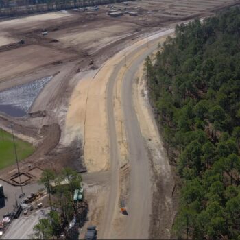 Aerial view: cleared site, dirt road, vehicles, pond, woods.