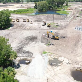 Aerial view: construction site with excavators, trucks, roads, greenery.