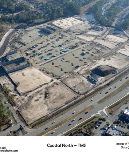 Aerial view: large cleared site, vehicles, near roads and homes.