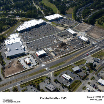 Aerial view of shopping center, parking lots, roads, nearby buildings.