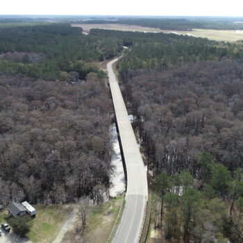 A straight bridge crosses forest, connecting roads; buildings visible.