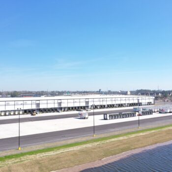 Warehouse with loading docks, trucks, grass, water, and clear sky.