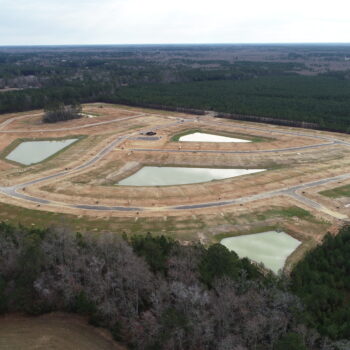 Aerial view: cleared site with roads, ponds, trees, surrounded by forest.