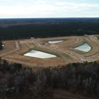 Aerial view: cleared construction site, ponds, roads, surrounded by forest.