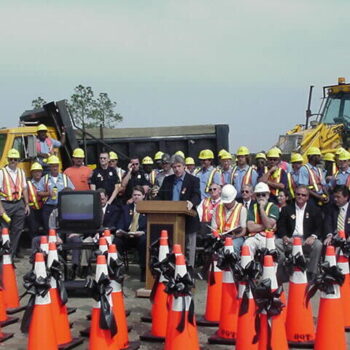 Workers and officials gather as man speaks by construction equipment.