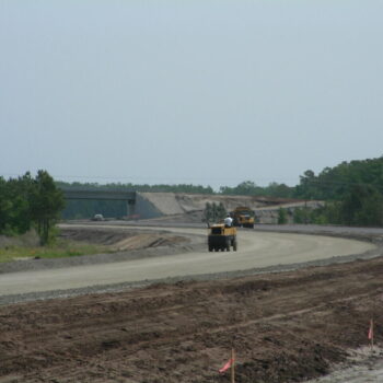 Construction vehicles on gravel road, trees and overpass visible.