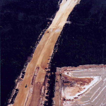 Aerial view: cleared strip, forest, vehicles, muddy foreground.