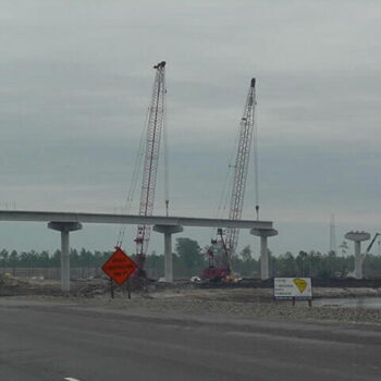 Highway under construction with cranes, supports, and work zone signs.
