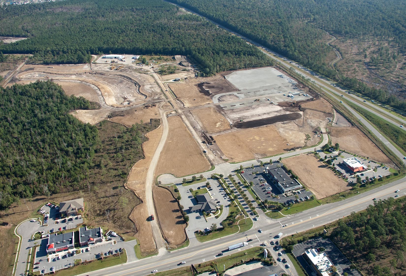 Aerial view: construction site, roads, buildings by highway and woods.