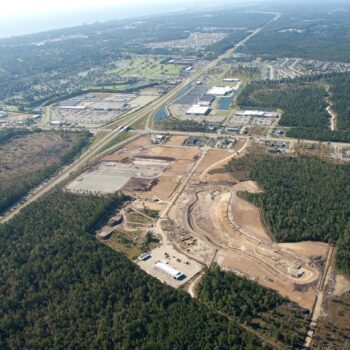 Aerial view: construction site amid forest, roads, and urban sprawl.