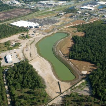 Aerial view: construction site, large pond, trees, buildings nearby.