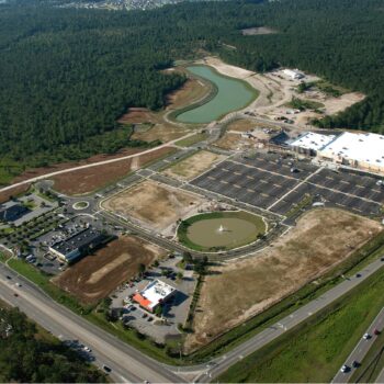 Aerial view: commercial area, large parking, pond, forest backdrop.