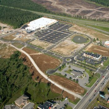 Aerial view: retail complex, parking, roads, roundabout, forest nearby.