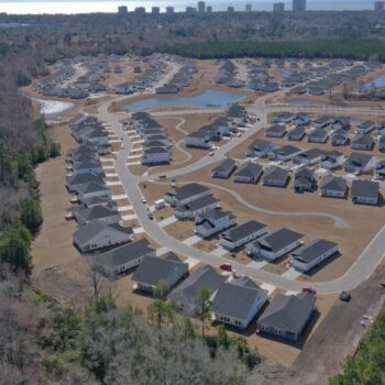 Aerial view: suburban homes, curving streets, pond, woods nearby.