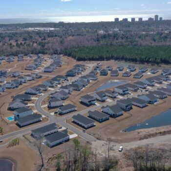 Aerial view: new suburban homes, roads, ponds, forest, city skyline.