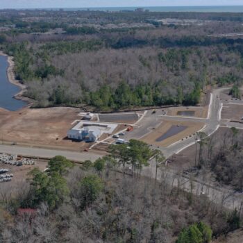 Aerial view: construction site, building, roads, woods, ocean nearby.