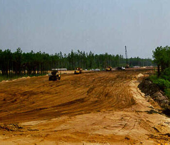 Construction vehicles on sandy site, trees and equipment in background.